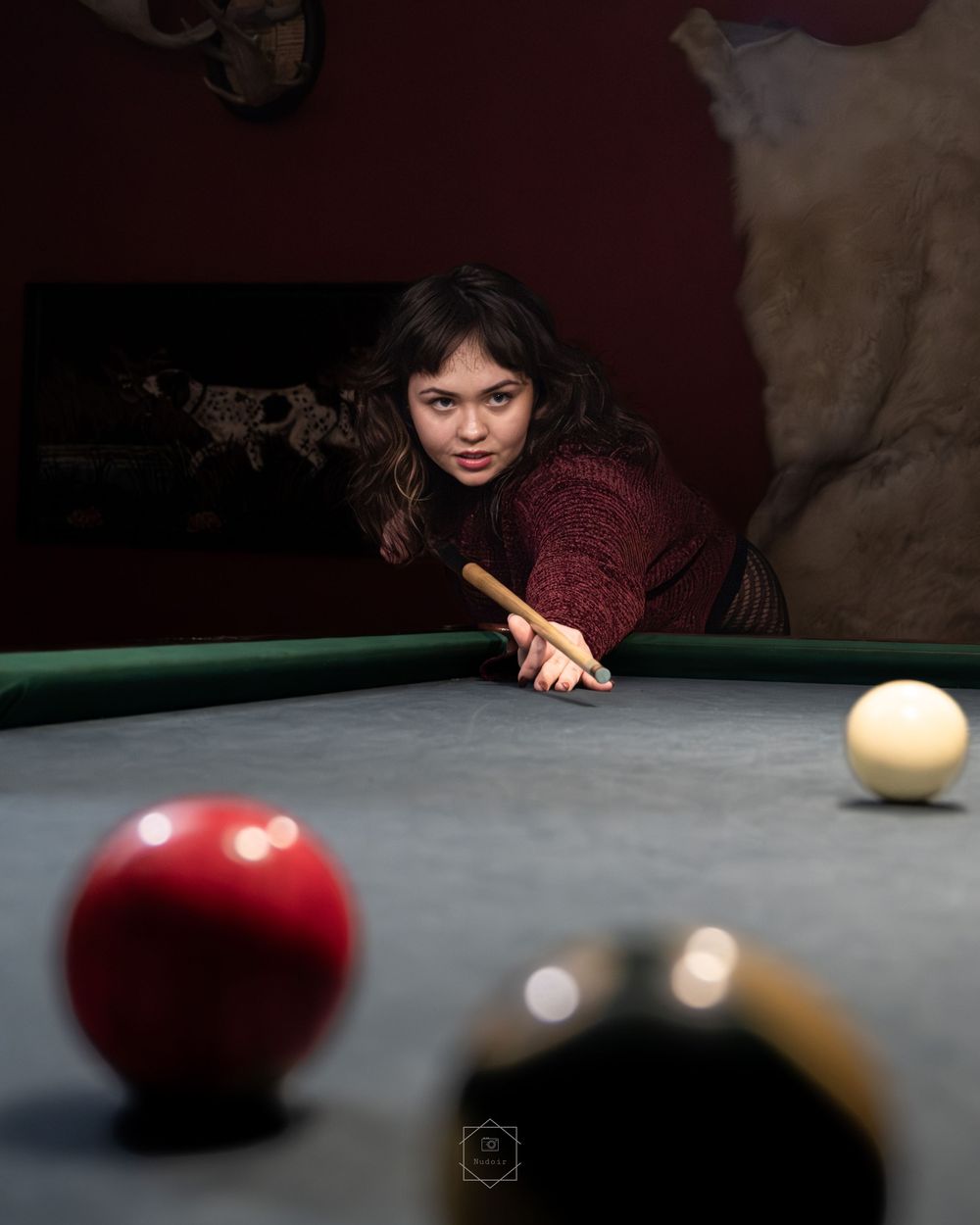 Young woman in red sweater leaning over a pool table, aiming a cue stick at billiard balls.