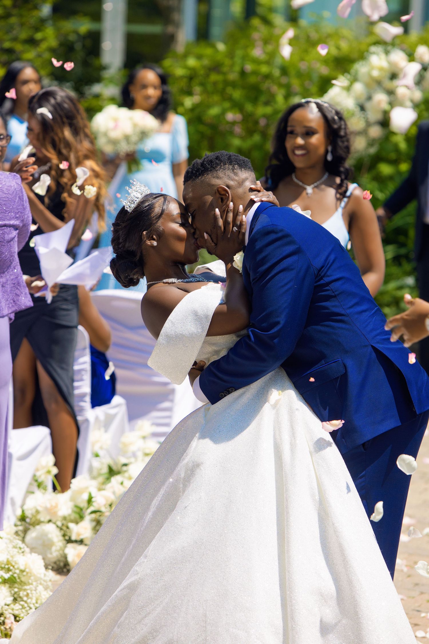 A joyful bride and groom share a romantic kiss outdoors at their wedding ceremony, surrounded by smiling bridesmaids in purple.