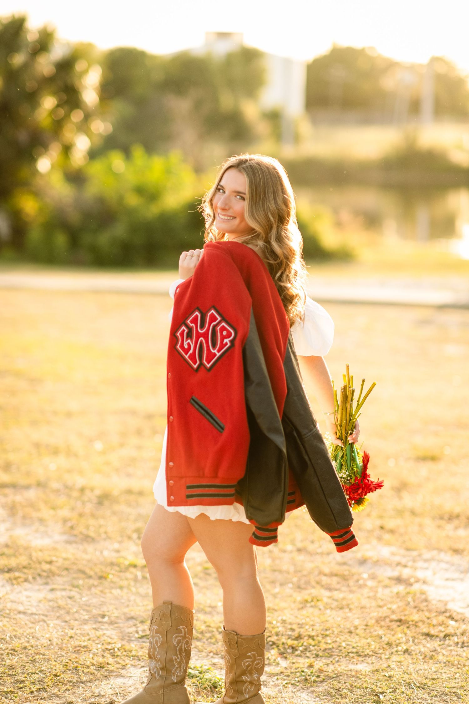 Smiling teen in red letterman jacket holding flowers, posing outdoors in golden hour light wearing cowboy boots.