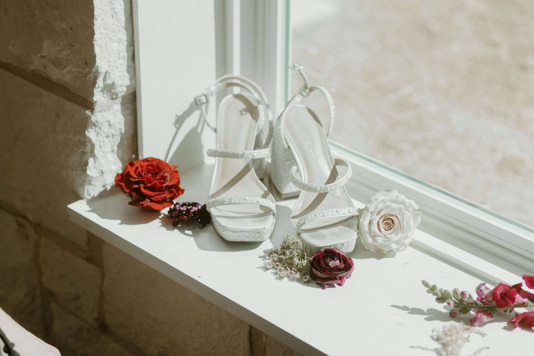 White bridal heels on windowsill surrounded by red and burgundy flowers for a wedding day detail shot.