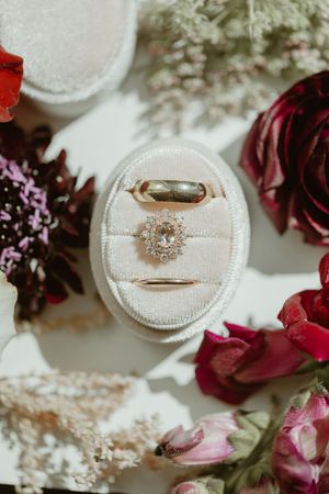 Elegant wedding rings displayed in a velvet oval box surrounded by red and pink florals.