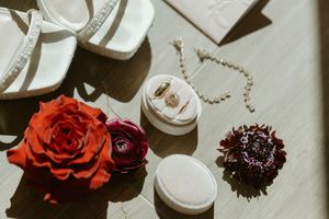 Wedding flat lay with white heels, red rose, ring box with gold bands, pearl necklace, and dark floral on wooden surface.
