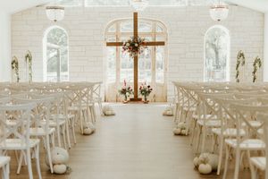 Elegant white chapel wedding venue with wooden cross, floral arrangements, white chairs, and pumpkin aisle decor.