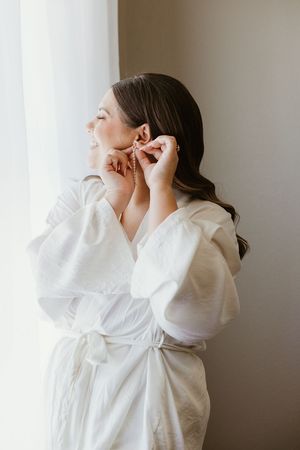 Bride in white robe putting on earrings by window light while getting ready on her wedding day.