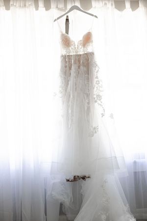 Elegant lace and tulle wedding dress hanging on a hanger in front of sheer white curtains.