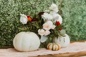 White pumpkins and floral arrangement with pink and red blooms on a rustic table against a green hedge backdrop.