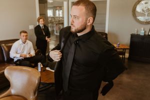 A groom in a black suit adjusts his tie while groomsmen prepare in the background before a wedding ceremony.