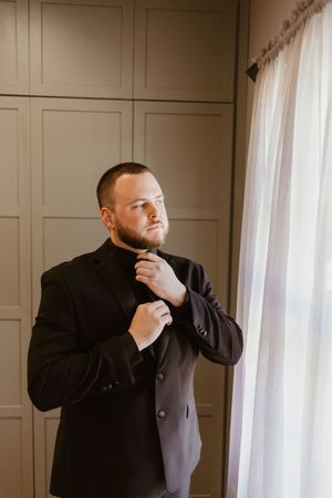 A well-dressed man in a black suit adjusts his tie near a bright window with sheer curtains.