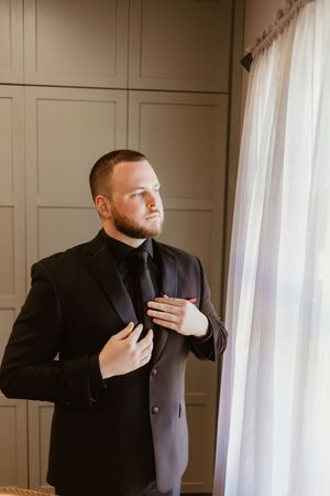 Groom in black tuxedo adjusting his lapel near a window with white curtains.