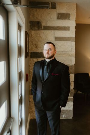 Groom in black tuxedo with red pocket square standing by stone wall and glass door.