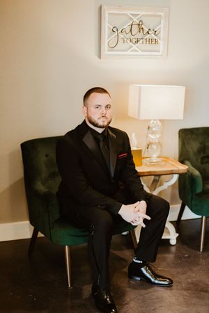 Groom in black tuxedo seated in green velvet chair beneath a gather together sign.