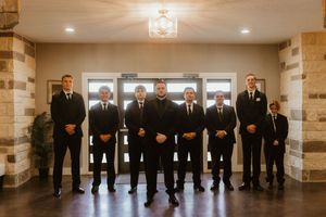 Groomsmen in black suits pose together in a elegant wedding venue lobby with stone walls and chandelier lighting.