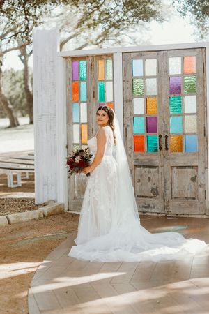 Bride in white gown with train poses before rustic doors with colorful stained glass panels outdoors.