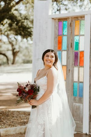 Smiling bride in lace wedding gown holding bouquet beside colorful stained glass doors outdoors.