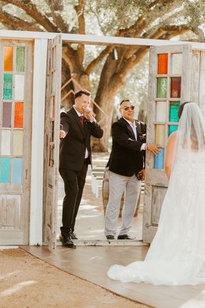 Emotional groom wiping tears as bride approaches colorful stained glass doors at outdoor wedding ceremony.