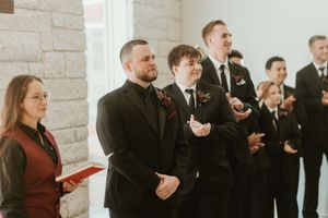 Groom in black tuxedo stands with groomsmen during wedding ceremony in white stone chapel.