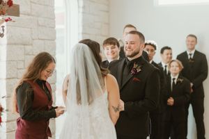 Groom smiles joyfully as bride faces him during vow exchange with officiant in burgundy dress.