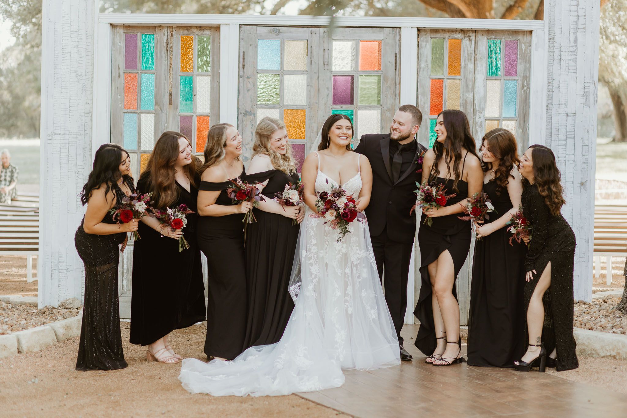 Bride and groom surrounded by bridesmaids in black gowns holding red bouquets at an outdoor wedding venue.