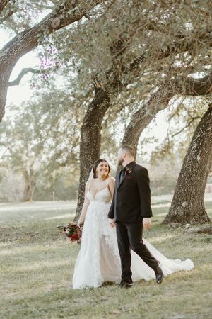 Bride and groom walk hand in hand under sprawling oak trees in an outdoor wedding portrait.