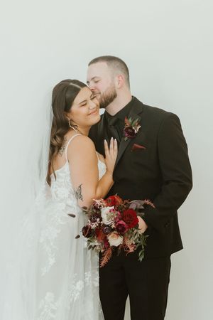 Bride smiles as groom kisses her cheek while holding a rich burgundy and red wedding bouquet.