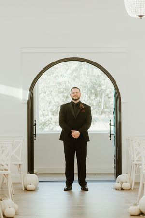 Groom in black tuxedo stands beneath arched doorway flanked by white pumpkins in bright chapel.
