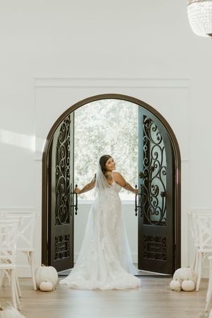 Elegant bride in lace gown stands in ornate arched iron doors of white wedding chapel interior.