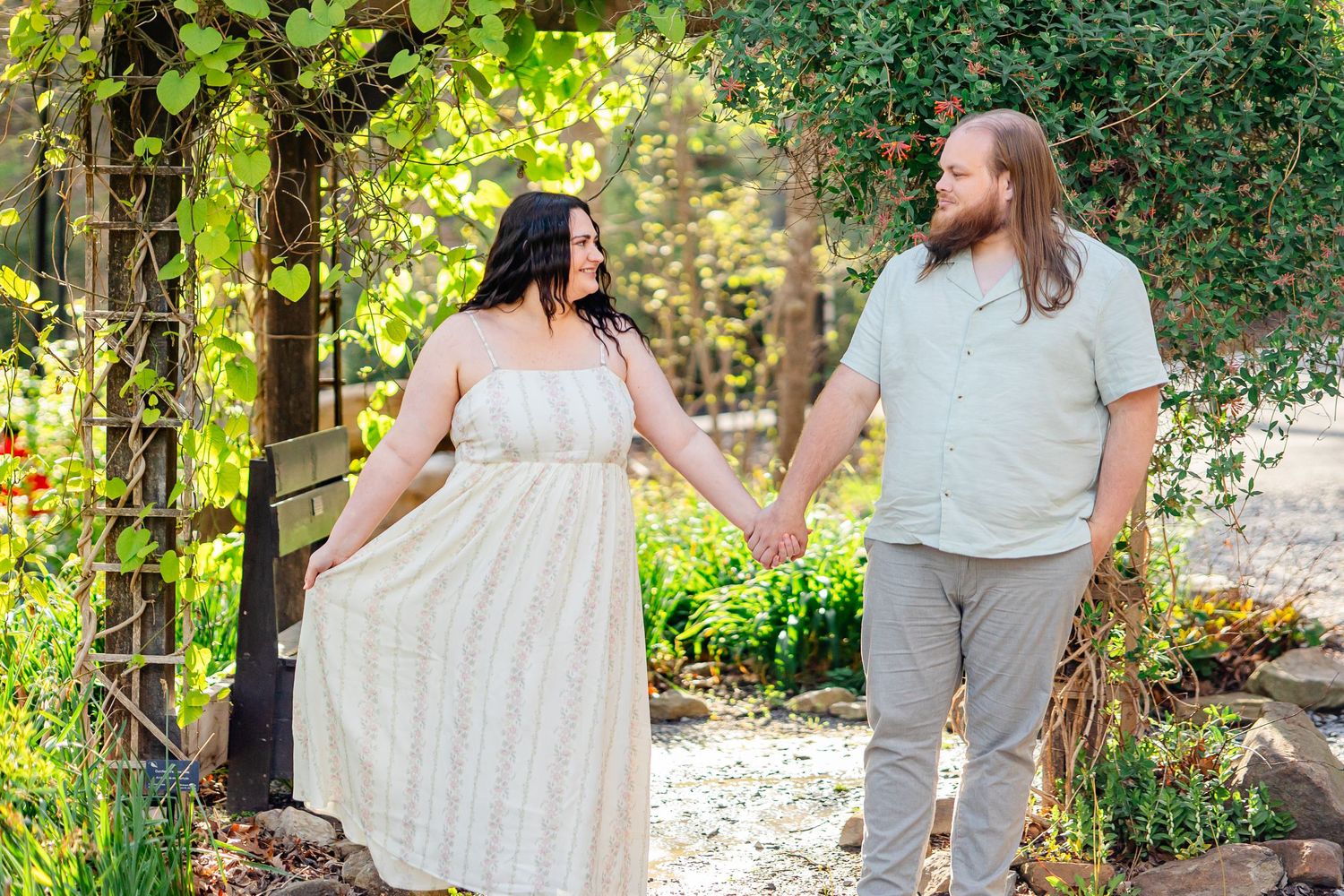 Couple holding hands in a lush garden under a vine-covered arbor, woman in white dress, man in light blue shirt and gray pants, morgantown