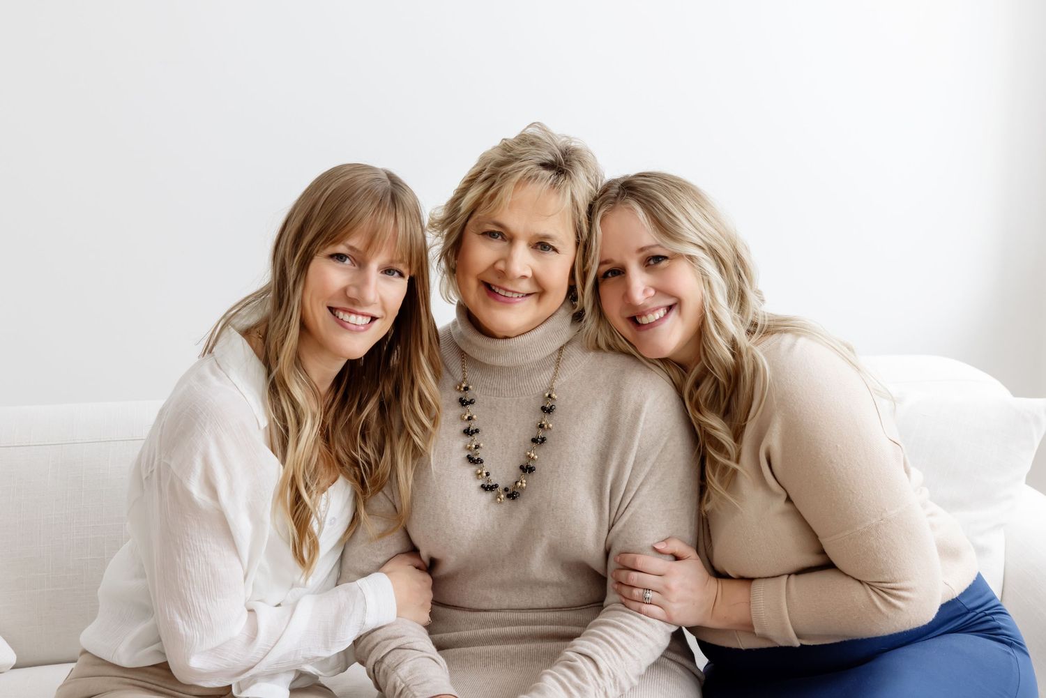 Three smiling women pose together on a white couch in a bright, elegant portrait setting.
