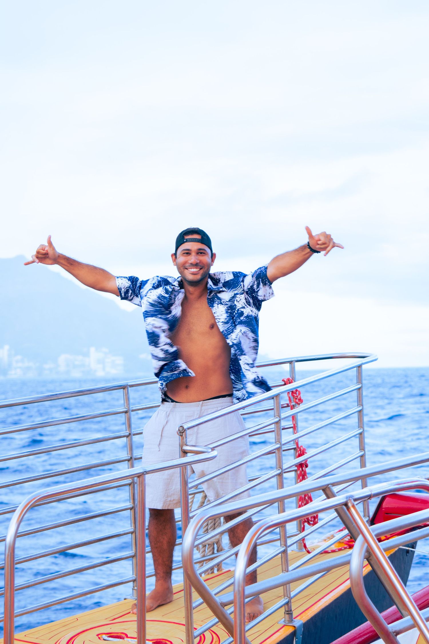 Man in open floral shirt and cap stands on boat deck with arms wide open, ocean and cloudy sky behind him.