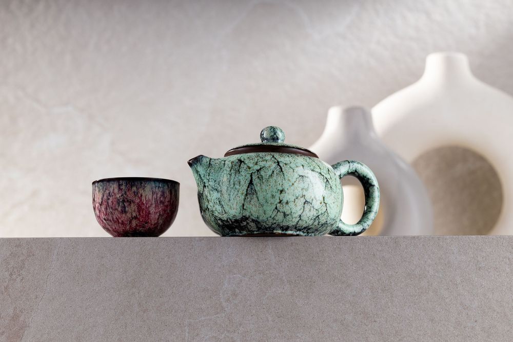 Handcrafted ceramic teapot with turquoise crackle glaze beside a red speckled tea cup and white vases on gray surface.