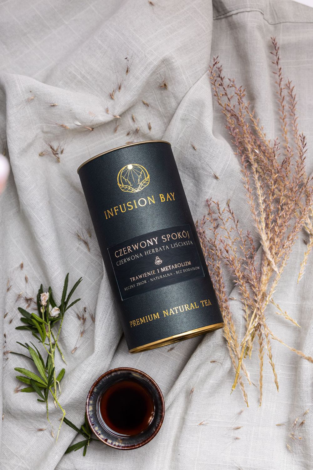 Infusion Bay Czerwony Spokój premium natural red tea canister with dried herbs and brewed tea cup on linen cloth.