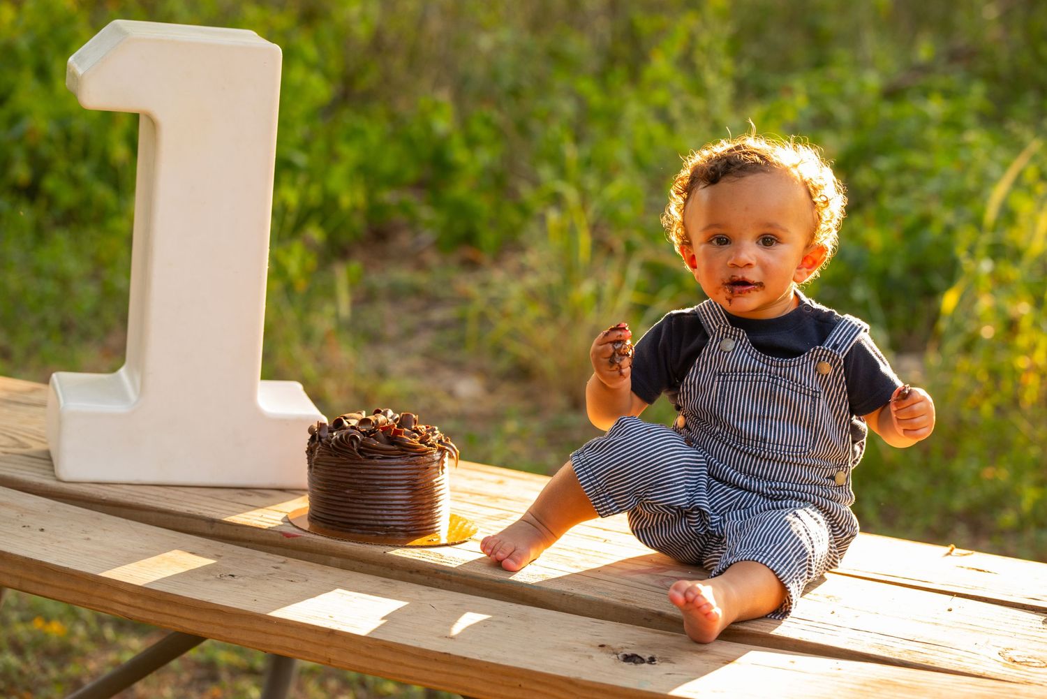 Baby boy celebrating first birthday sitting on wooden table with chocolate smash cake and large number one decoration outdoors.