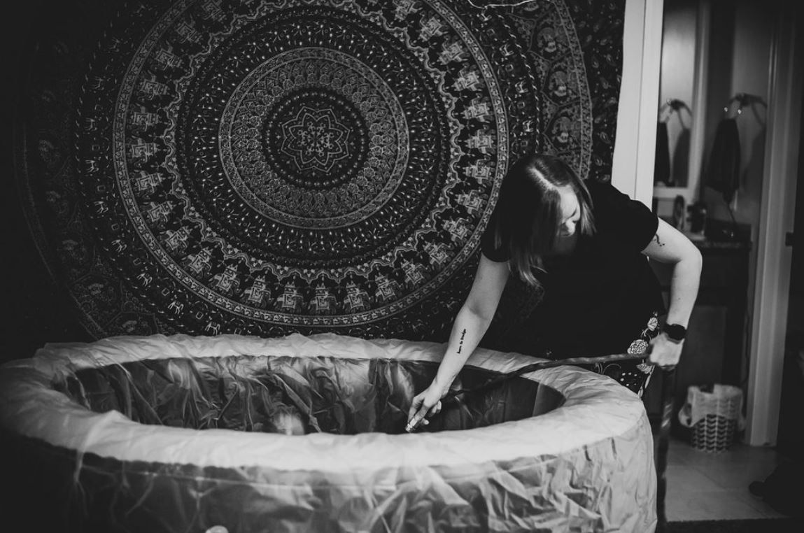 A black and white photo of a birthing pool with a mandala tapestry hanging on the wall.