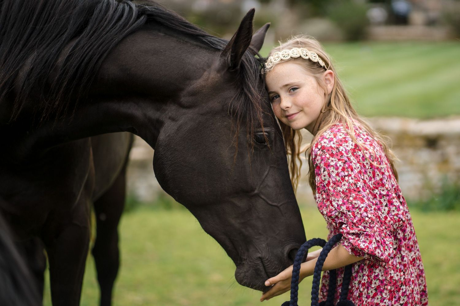Young blonde girl in floral dress tenderly embraces dark horse outdoors, eyes closed peacefully.