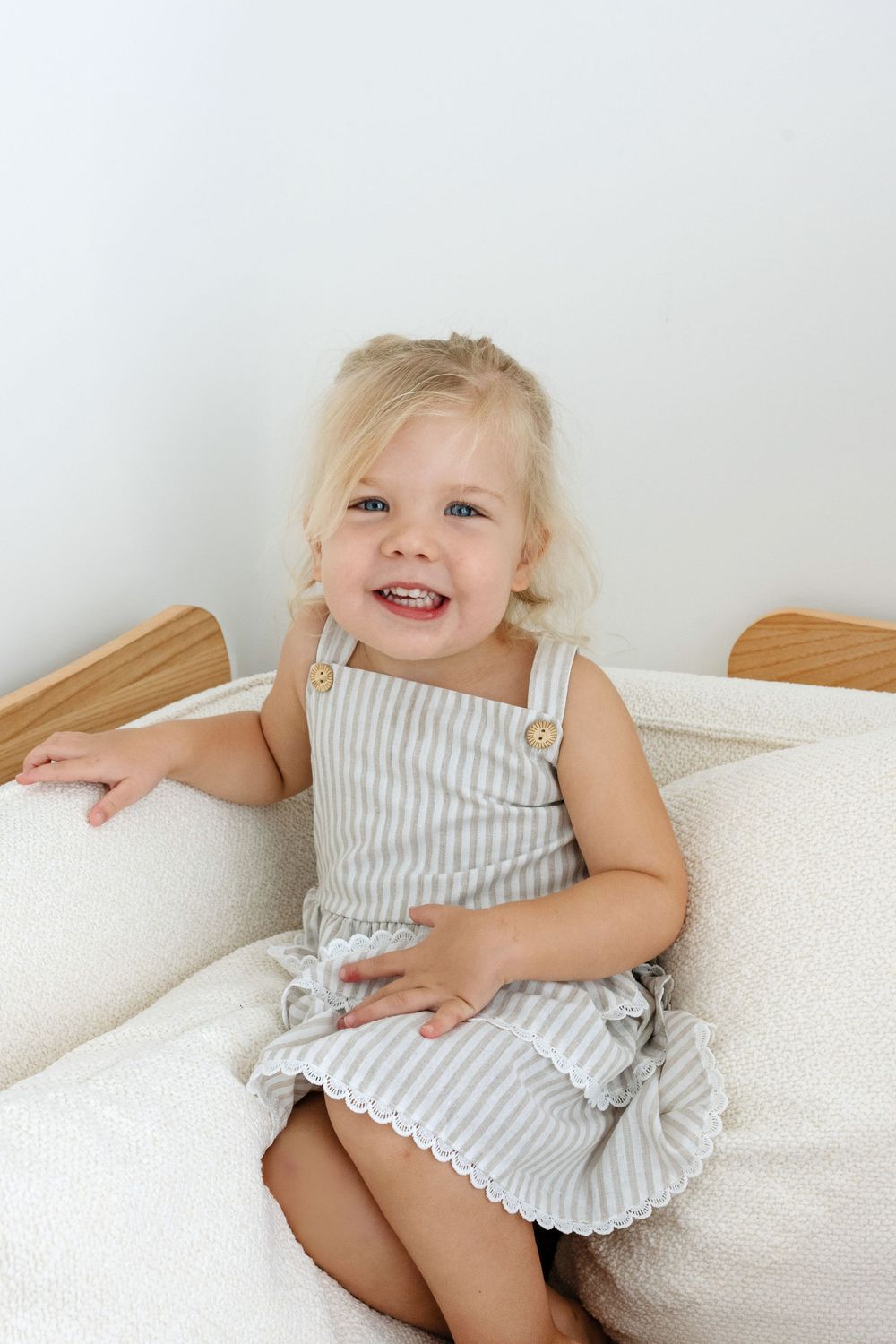 Smiling toddler girl with blonde hair sitting on a white sofa wearing a gray striped sundress with button straps.