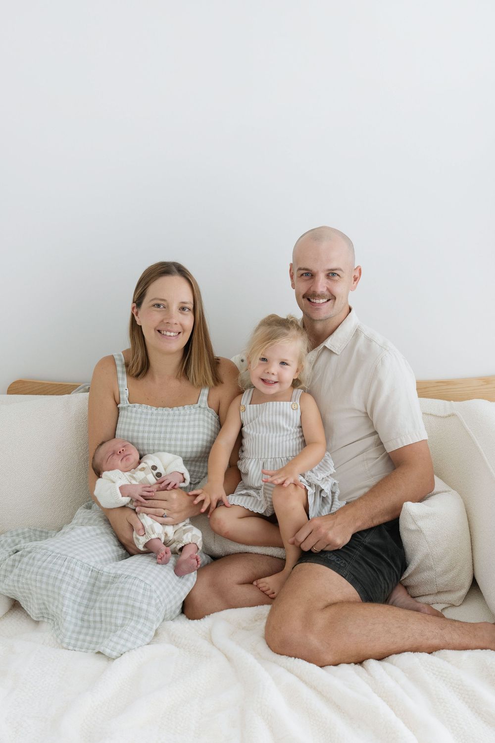 Happy family of four sitting on a white bed, mother holding newborn baby, toddler smiling, father embracing children.