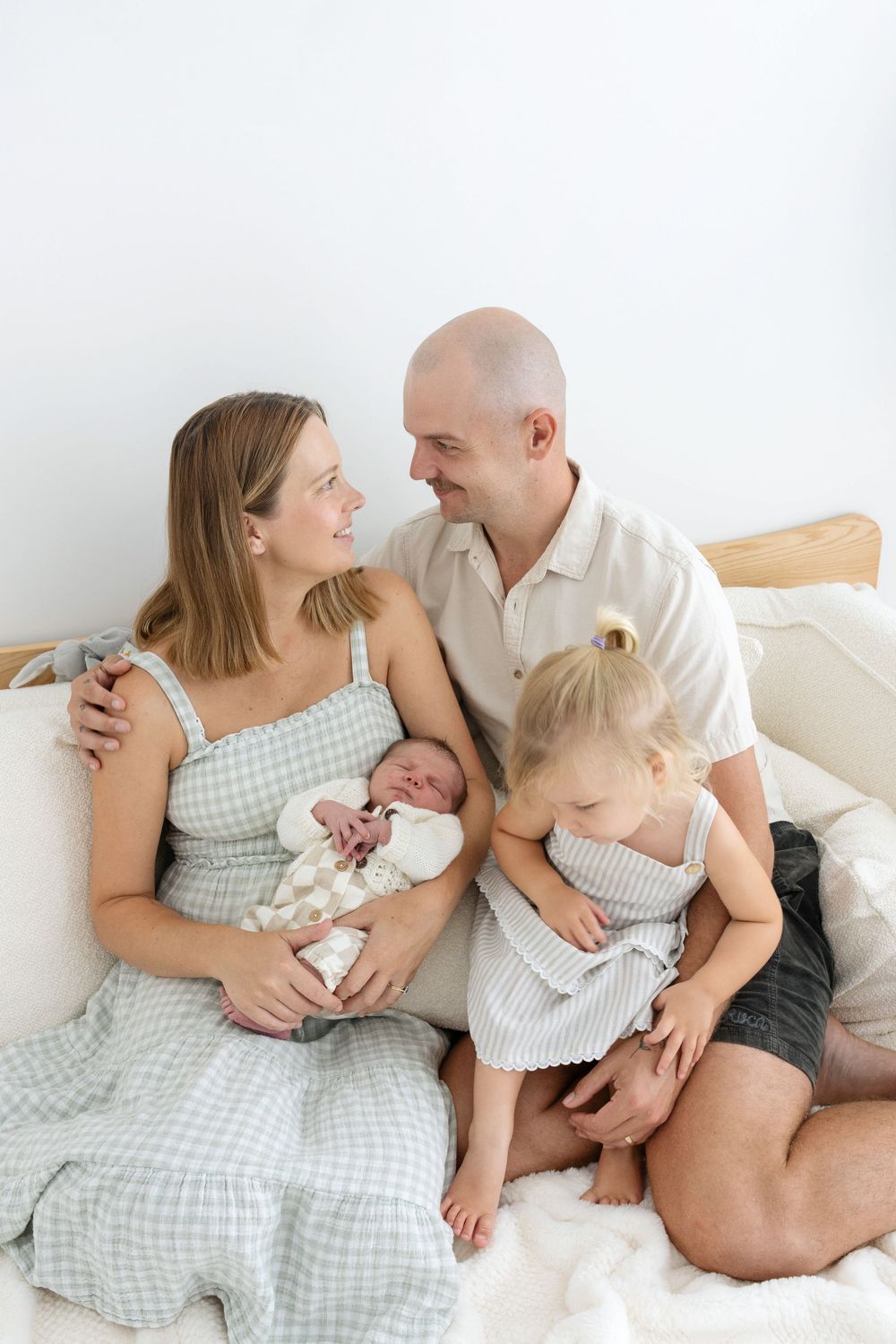 Happy family of four sitting on a white bed, parents smiling at each other while holding newborn baby and toddler daughter.