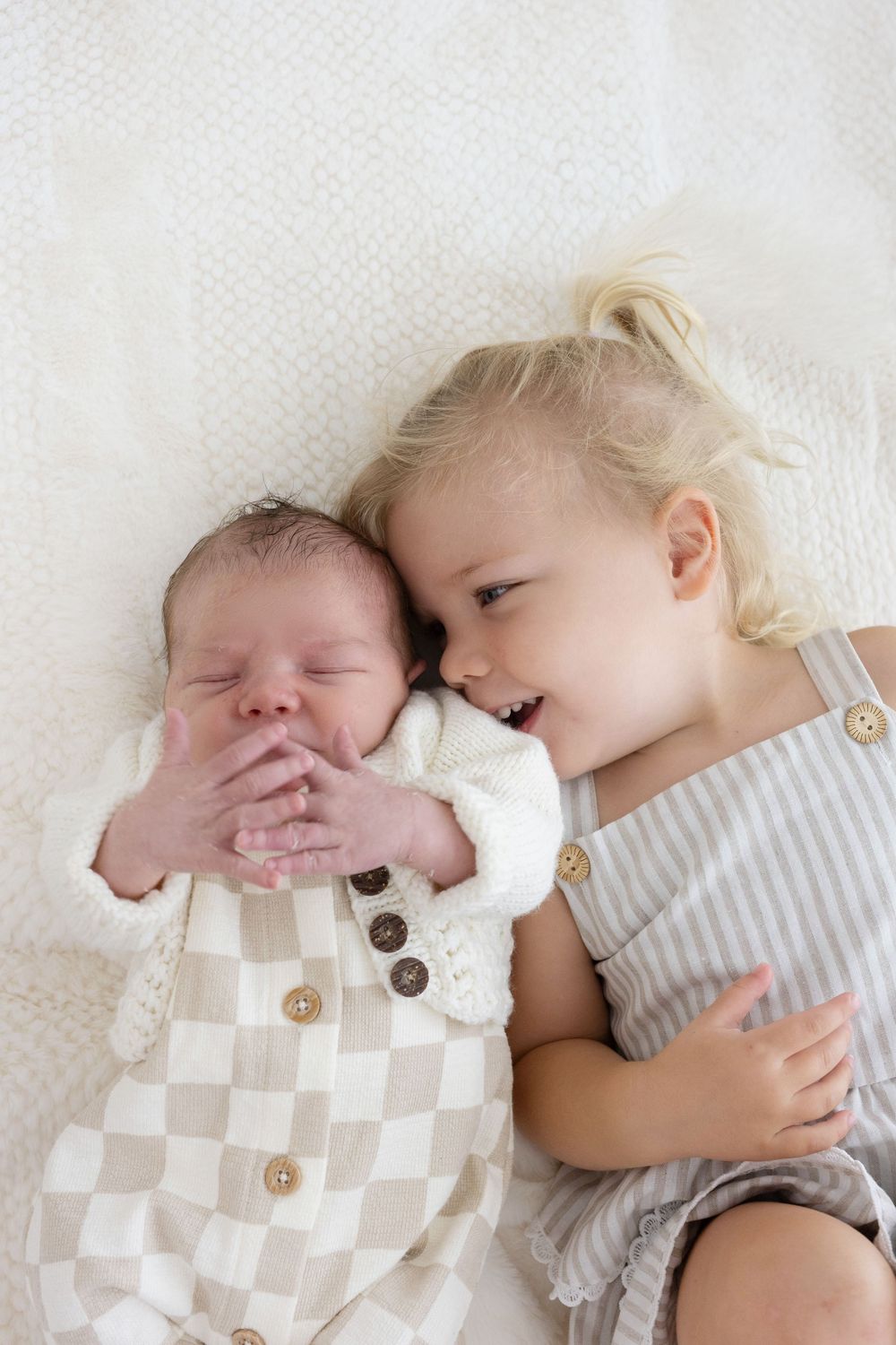 Overhead view of a blonde toddler girl snuggling cheek-to-cheek with her sleeping newborn sibling on a white blanket.
