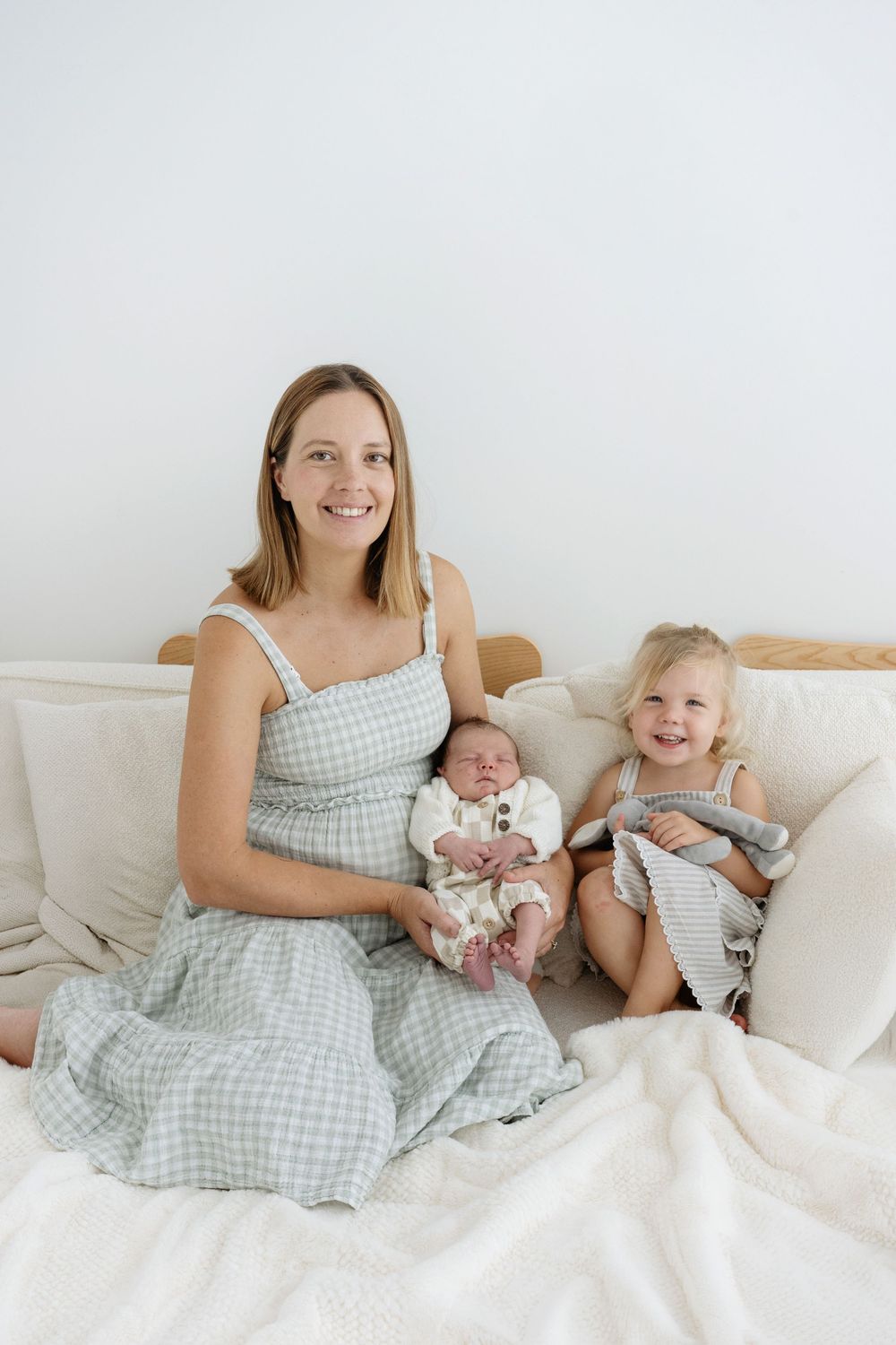 Smiling mother in gingham dress holding newborn baby beside toddler girl on white bed.