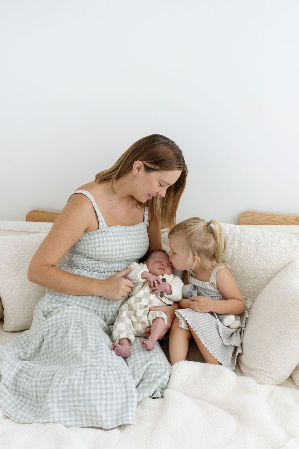 Mother in gingham dress sits on bed with toddler girl and newborn baby.