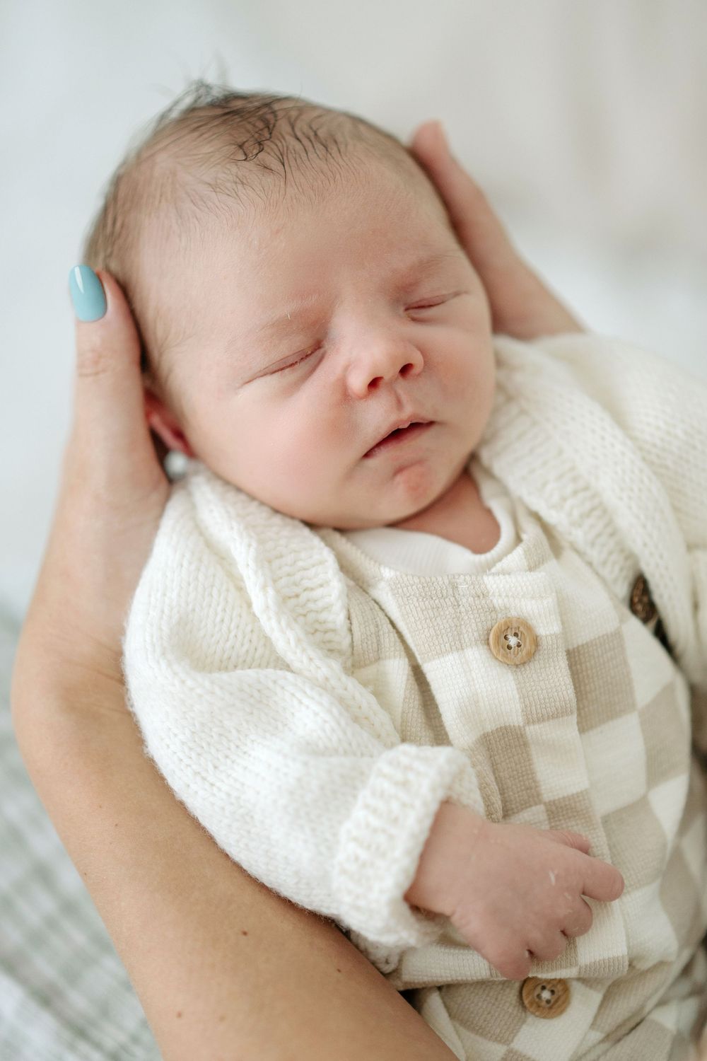 Newborn baby sleeping peacefully in adult hands, wearing a cream knit checkered outfit with wooden buttons.