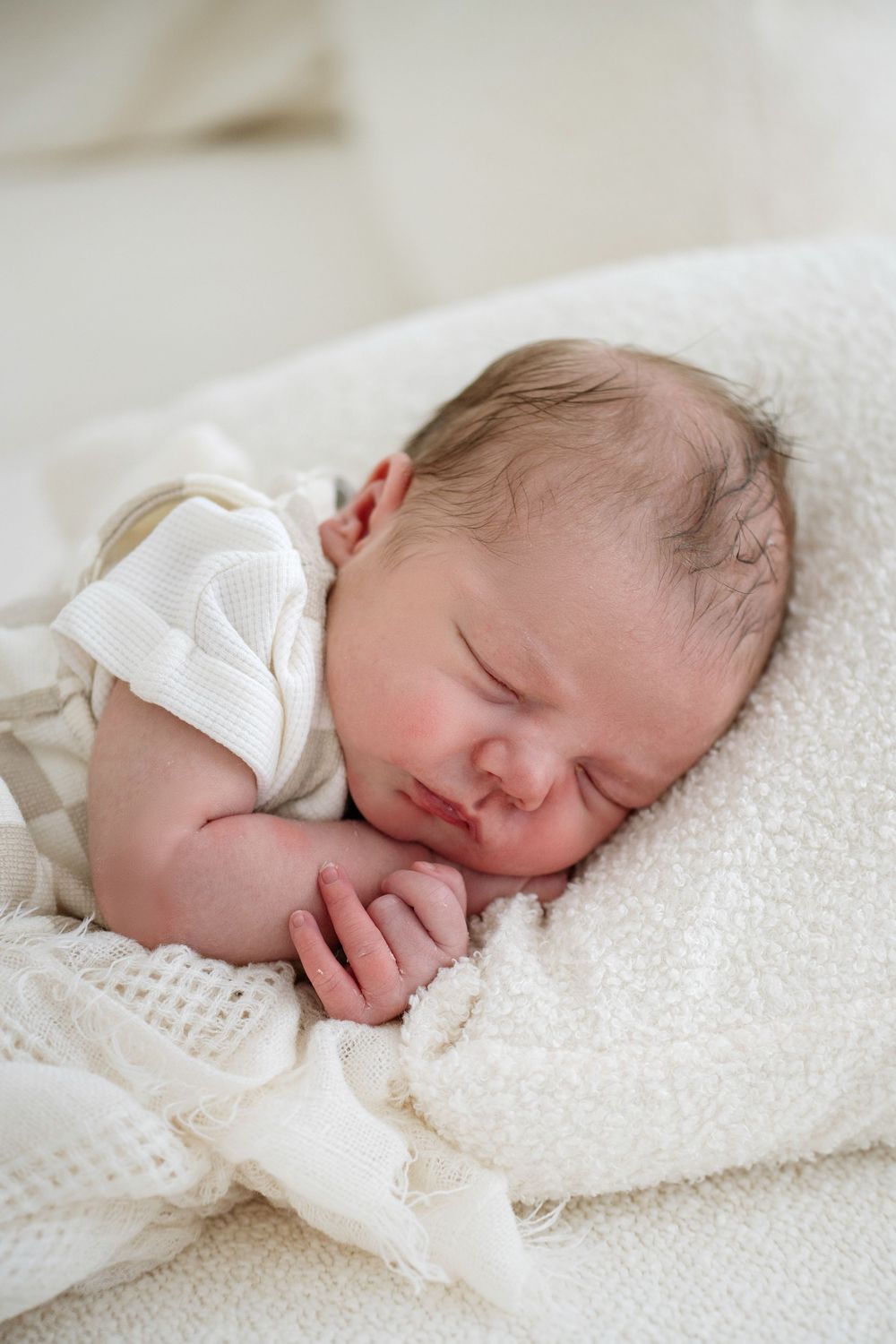 Peaceful newborn baby sleeping on white knit blanket wearing cream onesie with tiny hands tucked under chin.