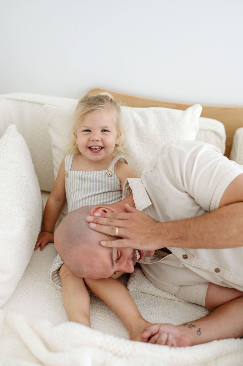 A laughing toddler girl sits on a bed with her bald father playfully nuzzling her lap in a bright, cozy room.