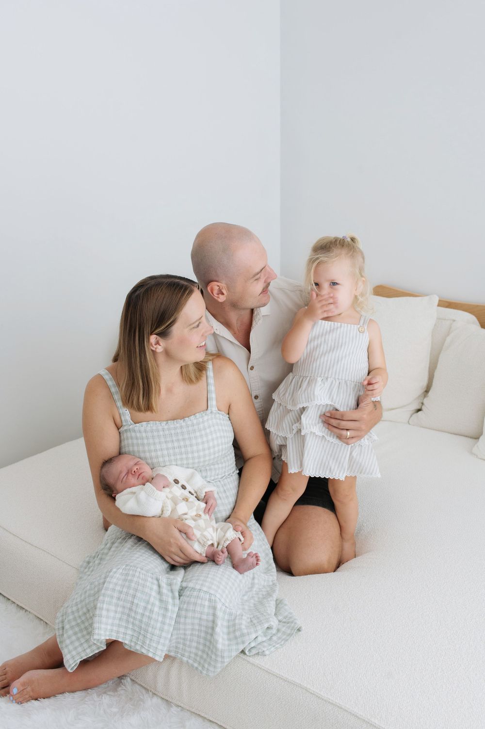 A family of four sits on a white couch, mom holding newborn, dad kissing toddler daughter in a bright studio.