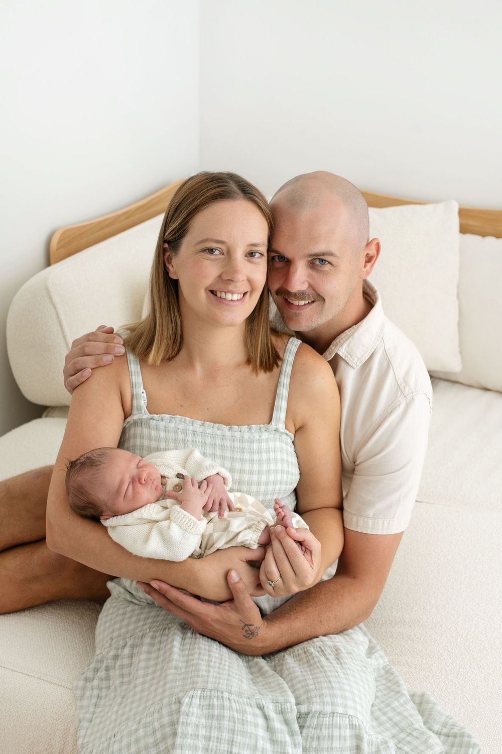 Smiling parents sitting on a cream bed holding their sleeping newborn baby wrapped in a white blanket.