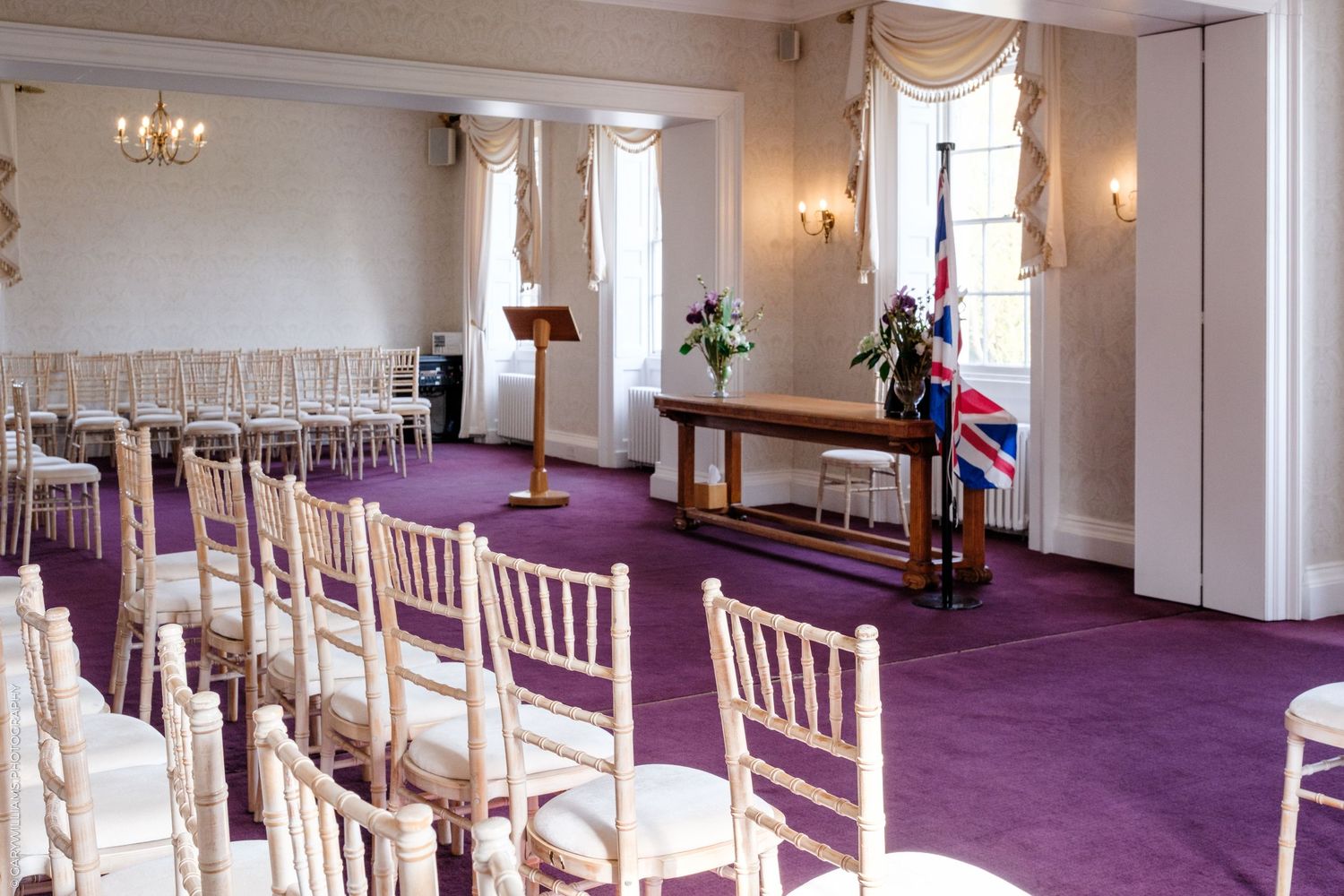 Elegant wedding ceremony room with white Chiavari chairs, purple carpet, wooden lectern, and Union Jack flag by tall windows.