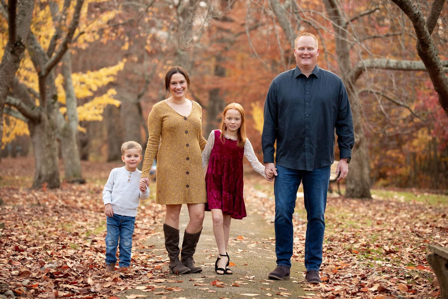 Family of four posing on a leaf-covered path surrounded by colorful autumn foliage trees.