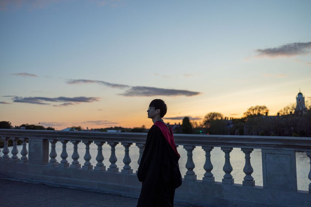 Harvard Graduate standing on John W. Weeks Bridge at sunset with a colorful sky, wearing a black coat with red accent.