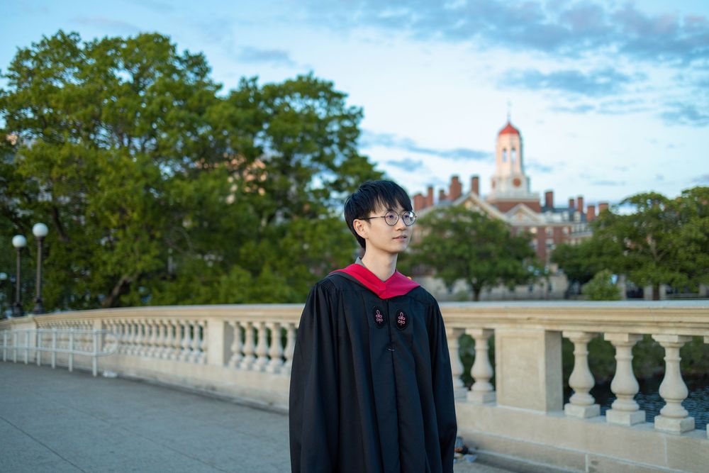 Harvard Graduate in black cap and gown with red hood poses outdoors on John W. Weeks Bridge
