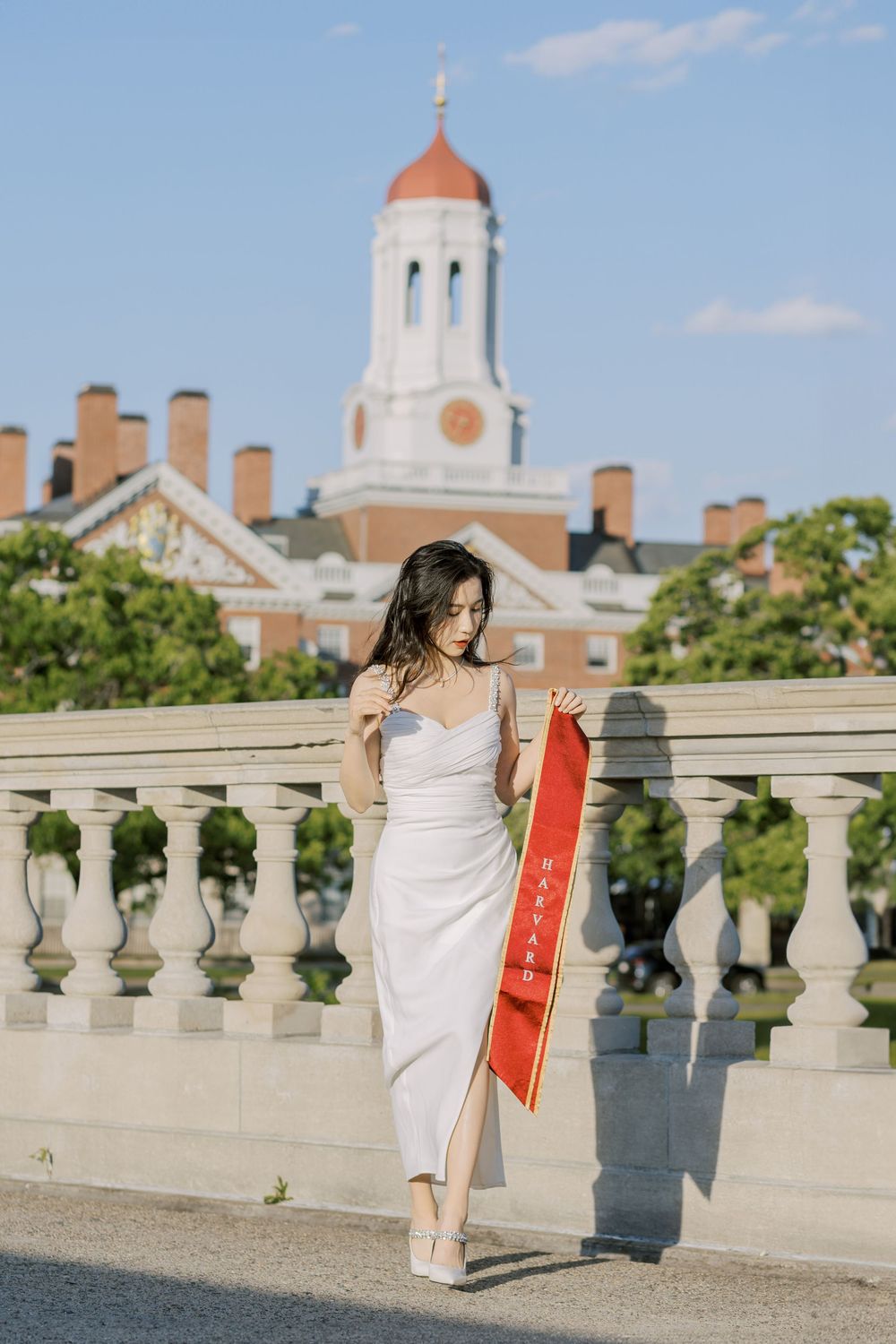 Harvard graduate in white dress holds red sash at historic campus bridge with iconic red-domed building backdrop.